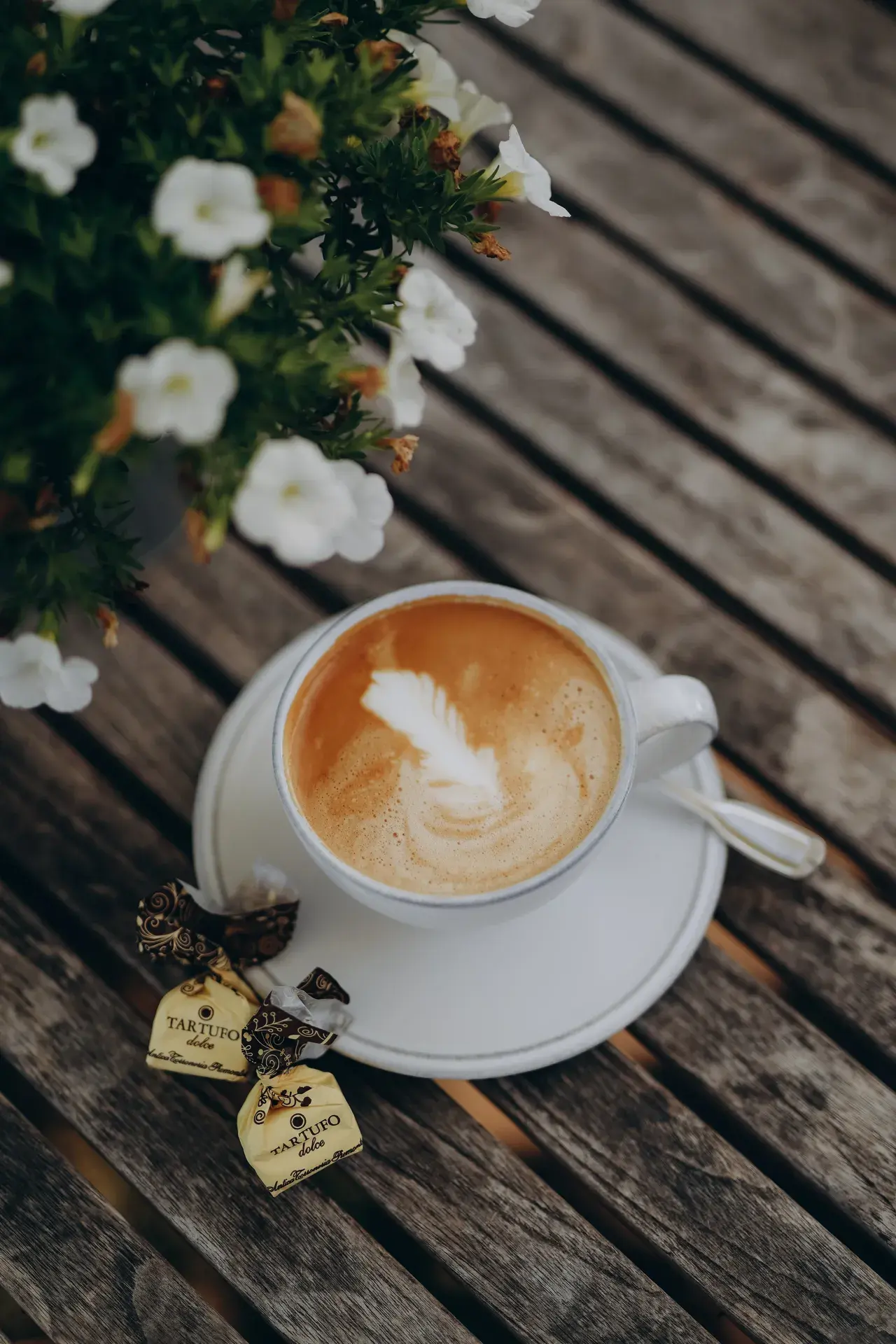 Cappuccino mit Milchschaum-Blatt in weißer Tasse auf einem Holztisch, daneben weiße Blüten